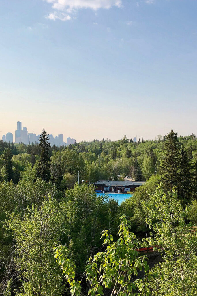 An aerial view of the Mill Creek Ravine, the Mill Creek pool surrounded by lush green forest, with a hazy downtown Edmonton in the background.