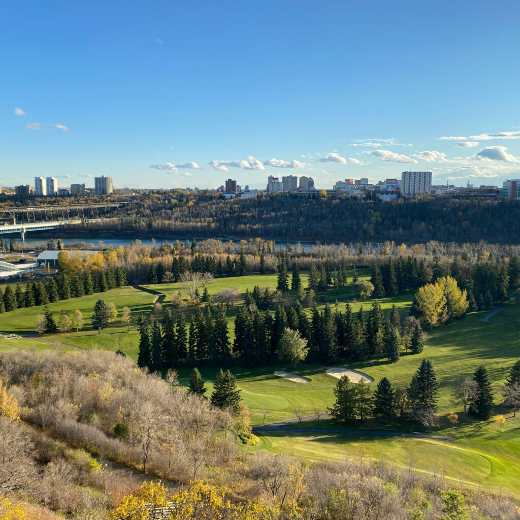 View over a green golf course in a river valley, with a river, bridge, and city skyline in the distance under a blue sky.