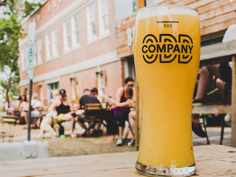 A mug of beer with the logo Odd Company sits on a table in the foreground, tables of people enjoy the patio in the background.