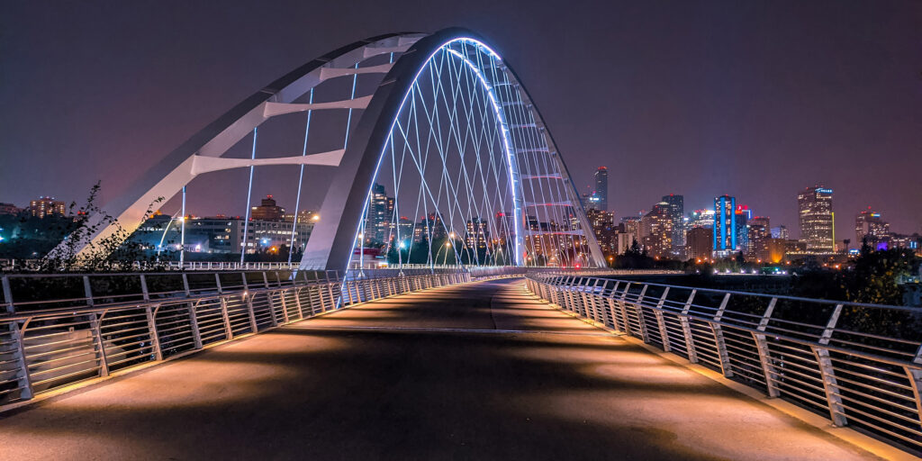 The Walterdale bridge lit up at night from a pedestrian view. Downtown Edmonton in the background.