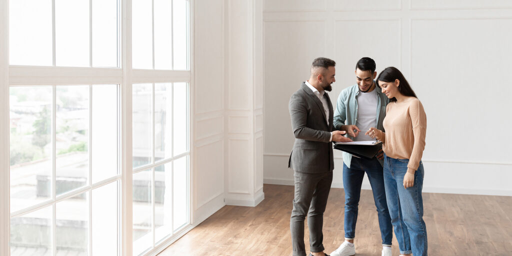 A REALTOR® meets with a couple in an empty apartment. They are referencing a clipboard at the centre of the image; the walls and floor are shades of beige.