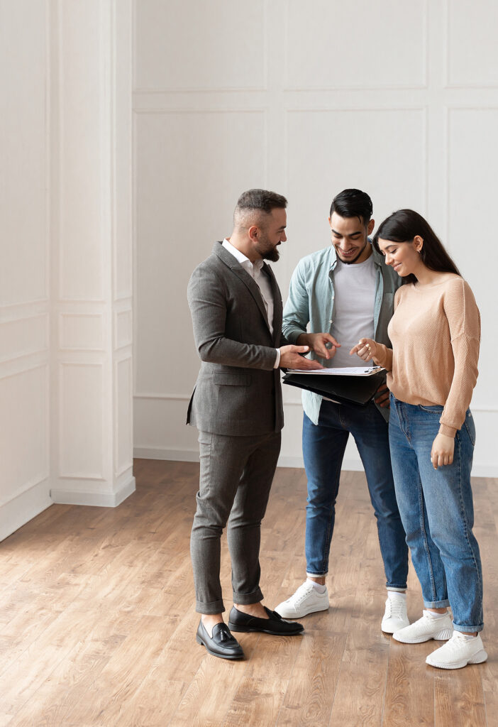 A REALTOR® meets with a couple in an empty apartment. They are referencing a clipboard at the centre of the image; the walls and floor are shades of beige.