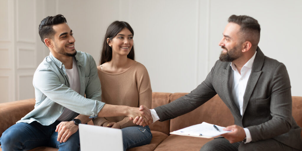 A real estate agent holding a clip board shakes hand with a young man, who is reaching across his partner.