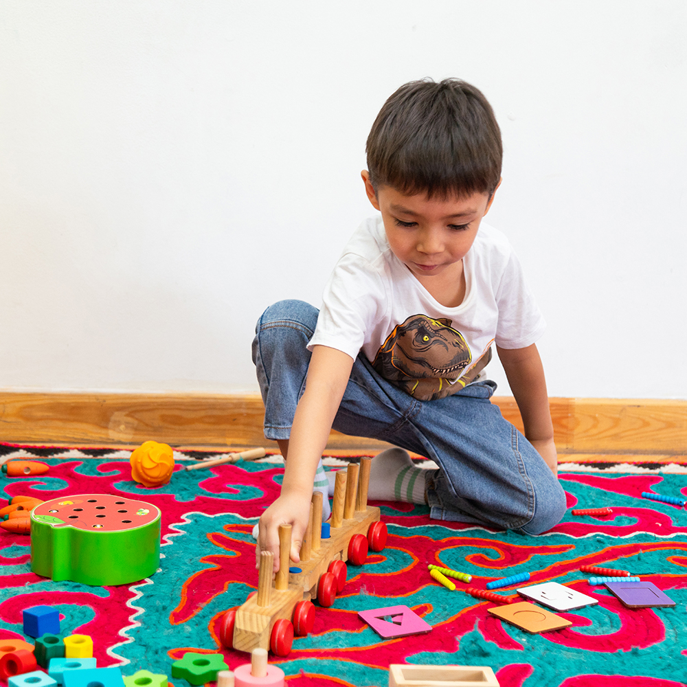 A boy playing with a wooden train on a turquoise and red rug.