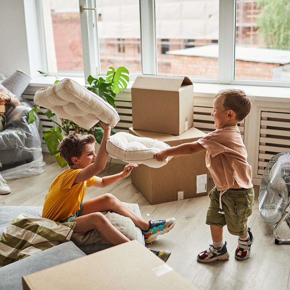 Two young boys have a pillow fight with couch cushions amid a moving scene.