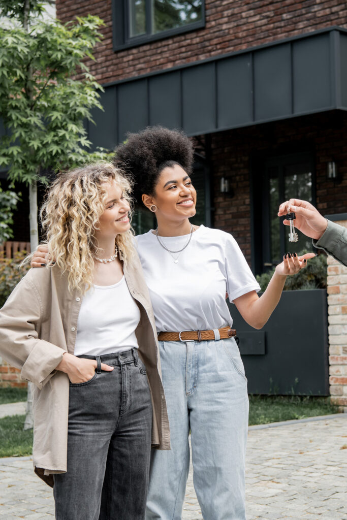 Successful REALTOR® passing a key for a new house to a pleased multiethnic lesbian couple.