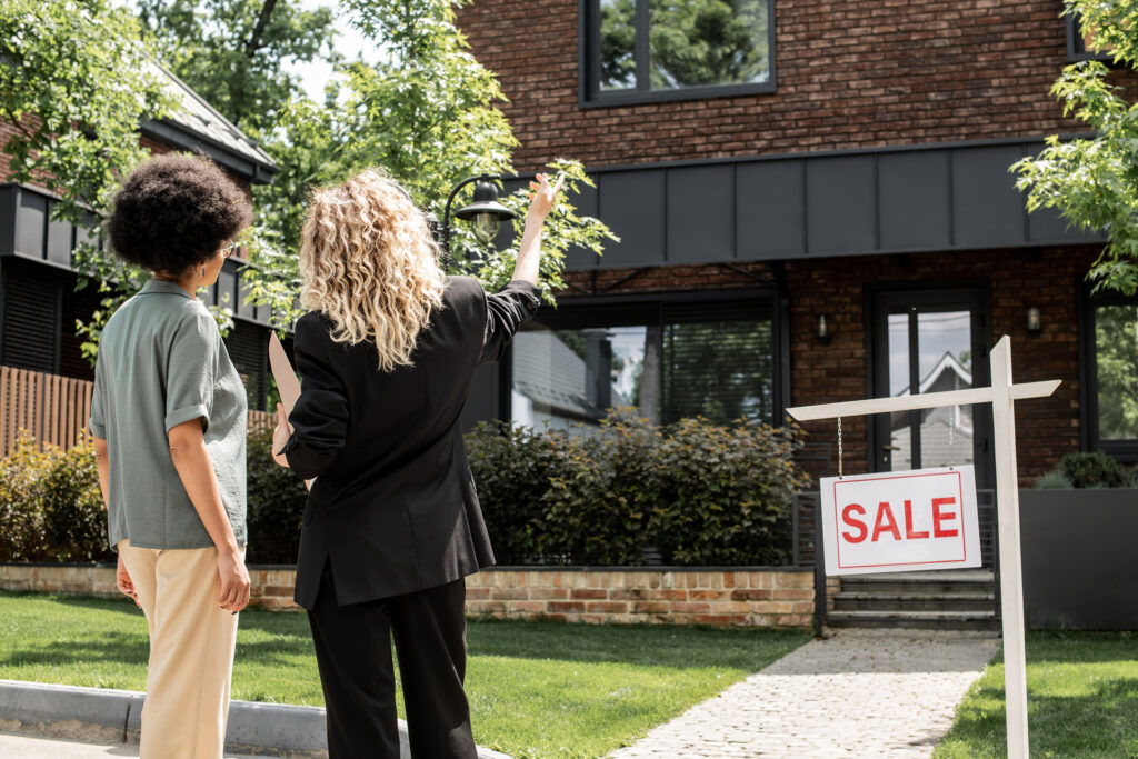 Two women face away from the camera, looking at property. One of the women gestures towards the home.