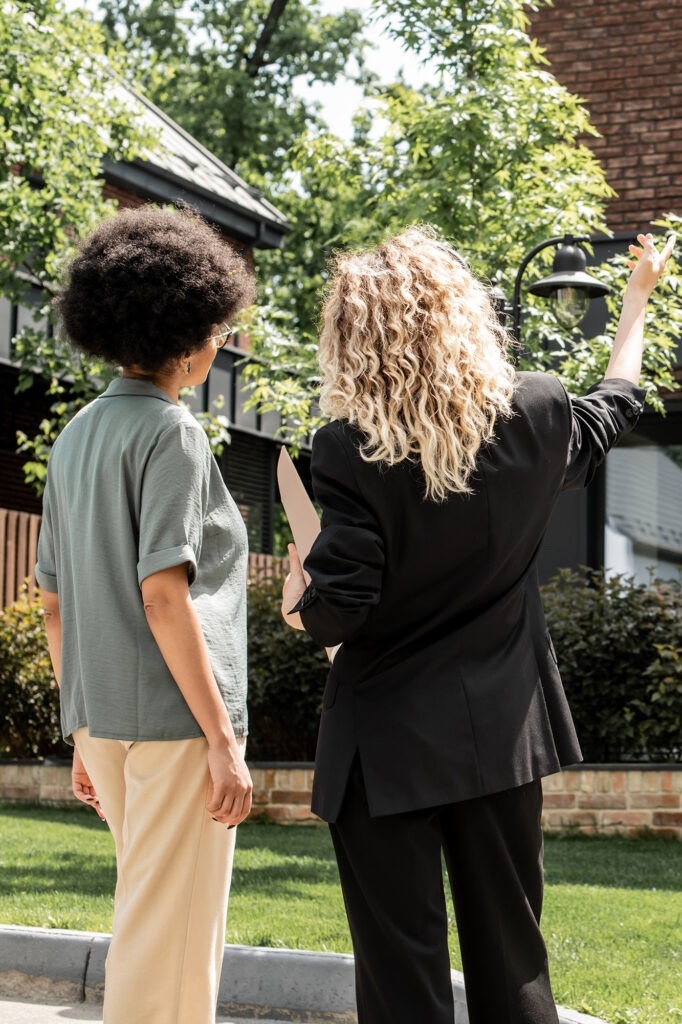 Two women face away from the camera, looking at property. One of the women gestures towards the home.